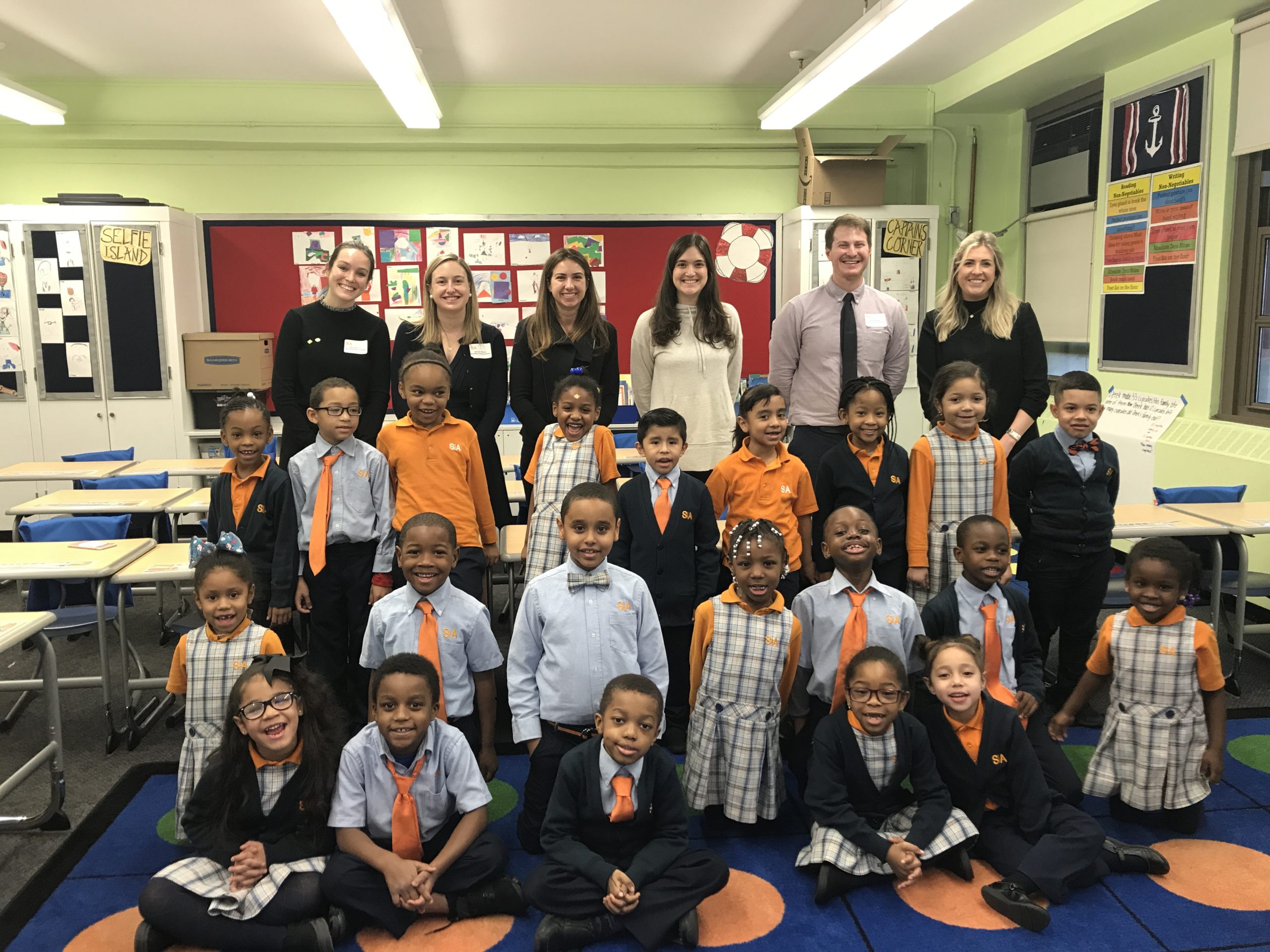 Children in uniforms pose smiling with teachers in classroom.